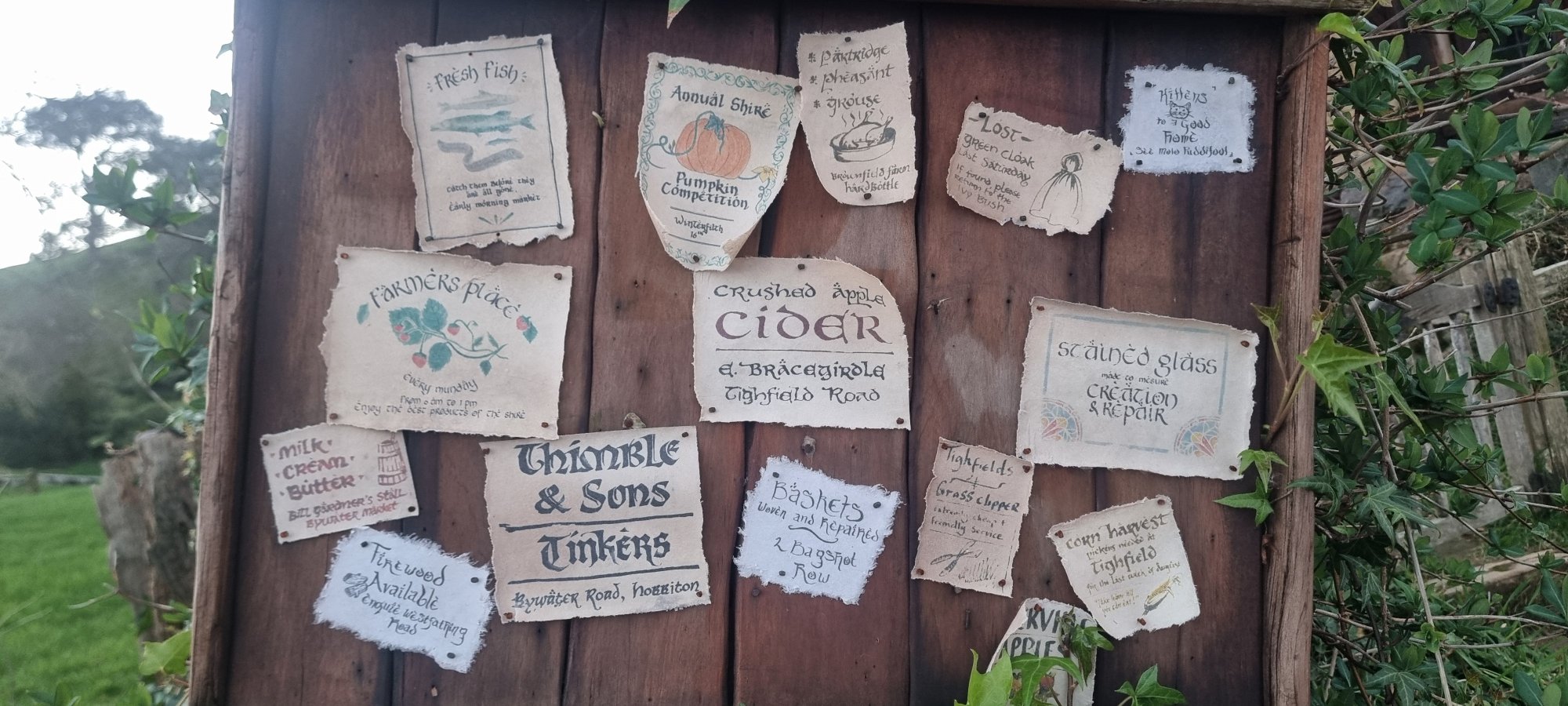 Hand-painted notice board at Hobbiton with Shire community notices for cider, fresh fish and tinkers