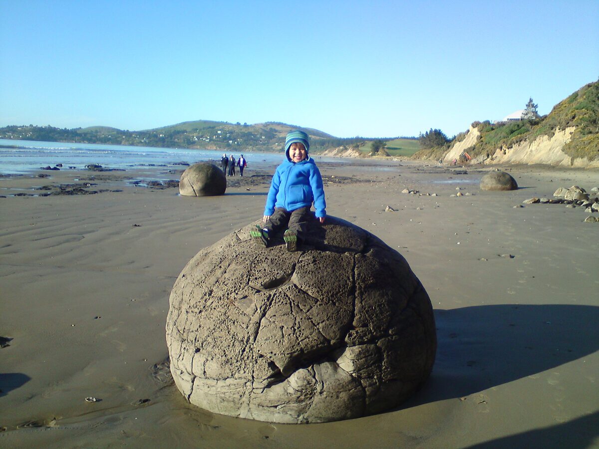 Child sitting on top of a Moeraki Boulder on Koekohe Beach with ocean and hills behind